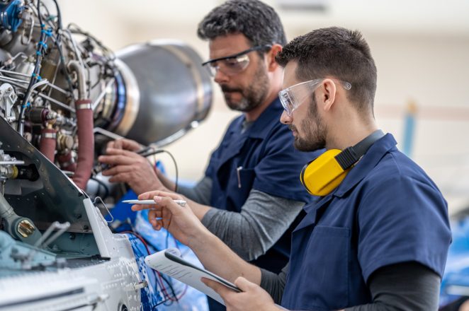 Aviation mechanics checking helicopter engine with multimeter, side view. Aircraft hangar workers in protective equipment repairing jet, medium shot